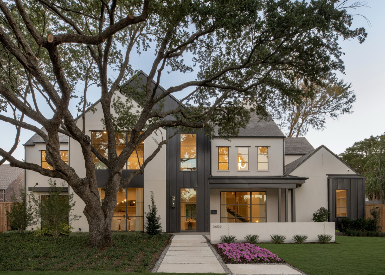 Modern white Dallas residence at twilight with full-height black steel windows glowing warm, framed by live oaks.