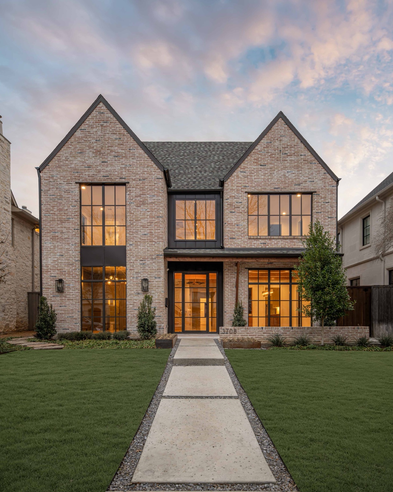 Light brick two-story home with tall black steel windows and a glowing steel entry door.